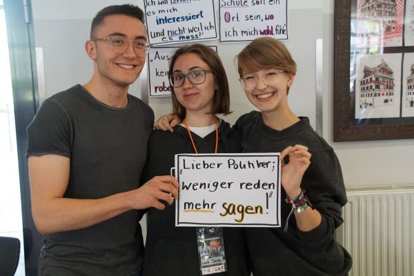 Drei Jugendliche halten ein Schild hoch mit der Aufschrift: Liebe Politiker; weniger Reden mehr Sagen"