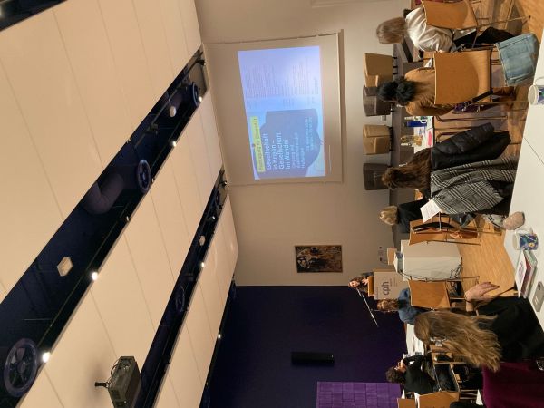 Foto in den Raum. Menschen von hinten sitzend. Zu sehen ist Marica Münch am Podium.