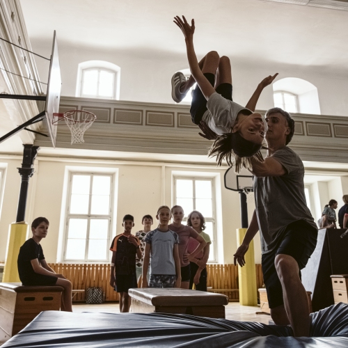 Jugendliche in einer Turnhalle. Ein Mädchen befindet sich gerade kopfüber im Sprung und wird beim Salto von einem Trainer unterstützt.