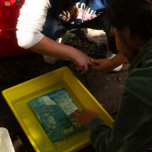 Ein Bild in blautönen befindet sich in einer Fotoentwicklungsschale mit der Flüßigkeit. Zwei Jugendliche hocken daneben und betrachten es gespannt.