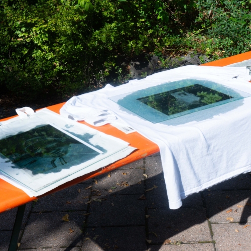 Weiße T-Shirts liegen auf einem Biertisch in der Sonne. Auf ihnen sind Schablonen und blaue Bilder zu erkennen, die in der Sonne trocknen.