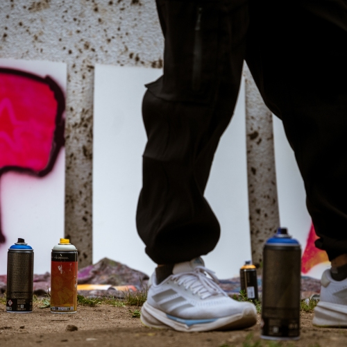 Das Bild ist auf Bodenhöhe fotografiert. Es zeigt Beine, sowie Spraydosen und im Hintergrund angefangene Spraybilder auf großen Platten.