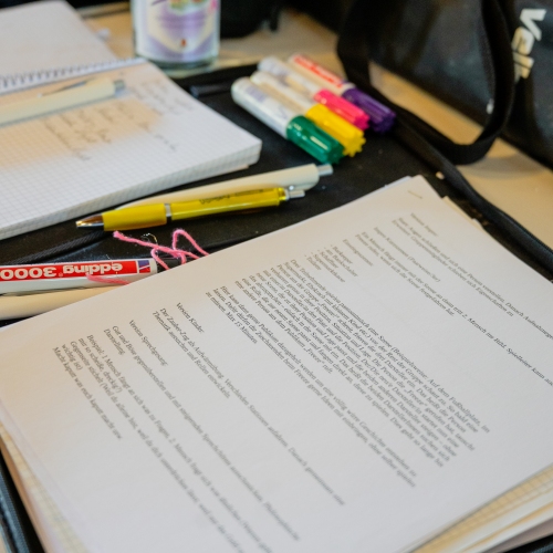 Eine offene Mappe liegt auf einem Tisch. Darin Stifte, Büroklammern und ein Skript mit Markierungen.