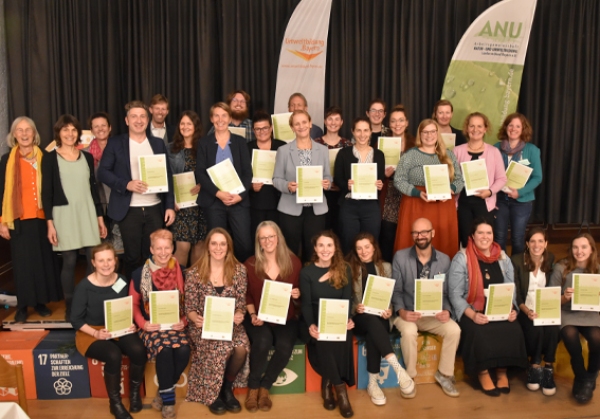 Die 26 Absolvent:innen der Weiterbildung Umweltbildung/BNE halten beim Gruppenfoto stolz ihre Abschlusszertifikate in die Kamera