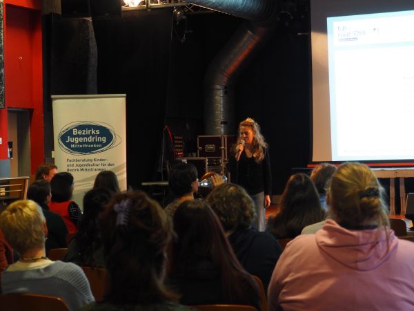 Begrüßung der Teilnehmenden, das Publikum ist von hinten zu sehen, die Moderatorin auf der Bühne