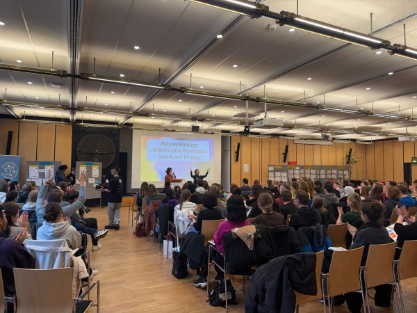 Blick von hinten in den Saal der Netzwerktagung. Voll besetzte Stuhlreihen.