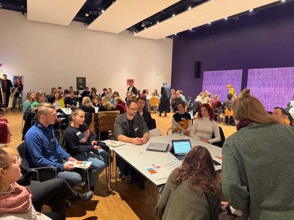 Foto der Netzwerktagung. Mehrere Sitzgruppen im Raum. Vorne im Bild eine Tischgruppe mit jungen Menschen und Erwachsenen, die sich austauschen.